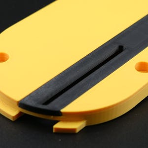 May include: A yellow and black 3D-printed object with a rounded shape. The object has a black stripe down the center and two circular holes. The object is likely a component or part of a larger item.