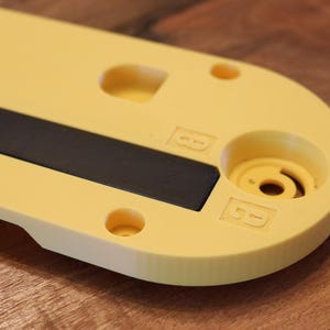 May include: A close-up of a yellow plastic object with a black rectangular insert. The object has rounded edges, several circular holes, and a cylindrical feature. The letters "B" are embossed on the surface. The background is a wooden surface.