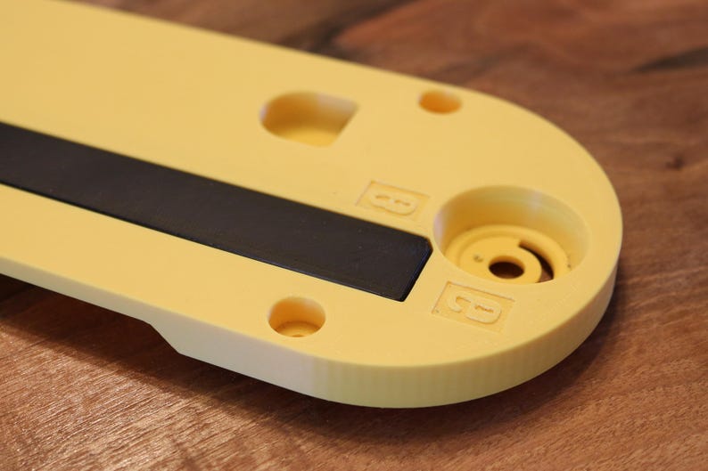 May include: A close-up of a yellow plastic object with a black rectangular insert. The object has rounded edges, several circular holes, and a cylindrical feature. The letters "B" are embossed on the surface. The background is a wooden surface.