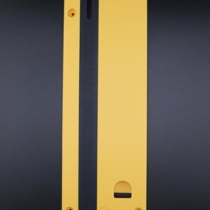 May include: A yellow table saw throat plate with a long, narrow black insert running vertically down the center. The plate features a rounded top, four screw holes, a small rectangular cutout, and a circular indentation with a lock icon.