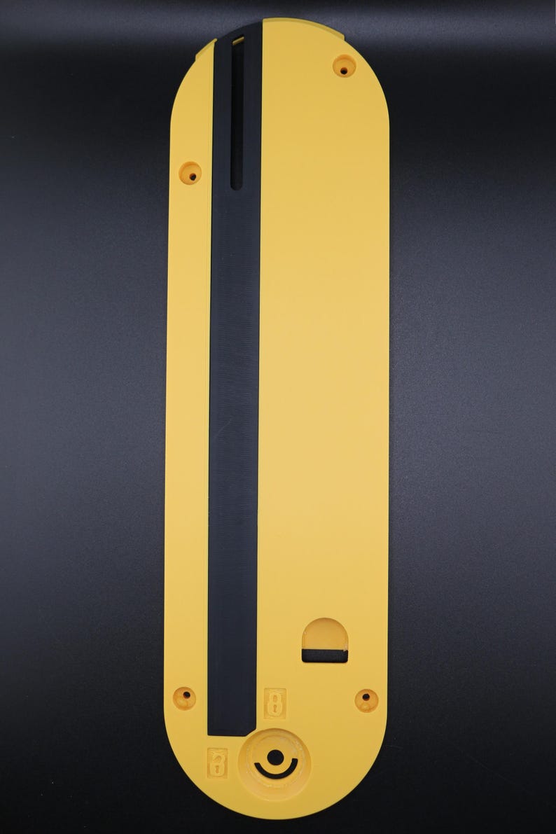 May include: A yellow table saw throat plate with a long, narrow black insert running vertically down the center. The plate features a rounded top, four screw holes, a small rectangular cutout, and a circular indentation with a lock icon.
