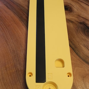 May include: A yellow and black plastic component with a rounded end and a black stripe down the center. The component has several holes and a circular indentation with a smiley face design.