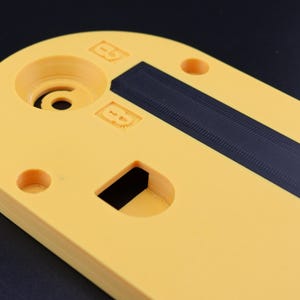 May include: A yellow, oval-shaped plastic component with a black, textured strip. The component has several circular holes and a rectangular cutout. The surface has embossed markings and a central circular feature.
