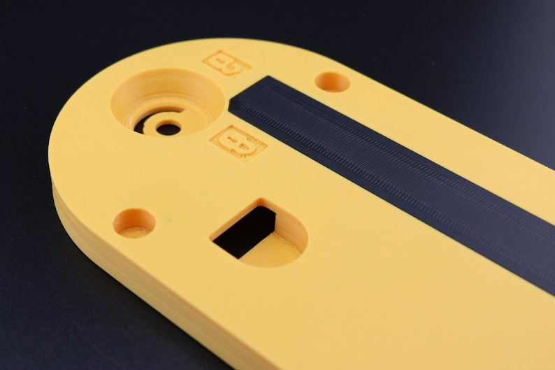 May include: A yellow, oval-shaped plastic component with a black, textured strip. The component has several circular holes and a rectangular cutout. The surface has embossed markings and a central circular feature.