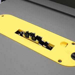 May include: A yellow table saw blade guard with a black circular blade and a gray table surface. The guard has a rectangular opening exposing the saw blade teeth. The guard is secured with screws and a black knob.