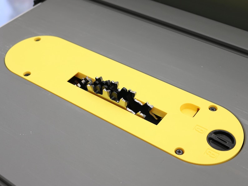 May include: A yellow table saw blade guard with a black circular blade and a gray table surface. The guard has a rectangular opening exposing the saw blade teeth. The guard is secured with screws and a black knob.