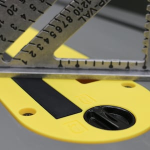 May include: A silver metal rafter square with black markings rests on a yellow circular table saw insert plate. The plate features a black rectangular slot and a black circular locking knob. The square displays the text 100th and PIVOT.