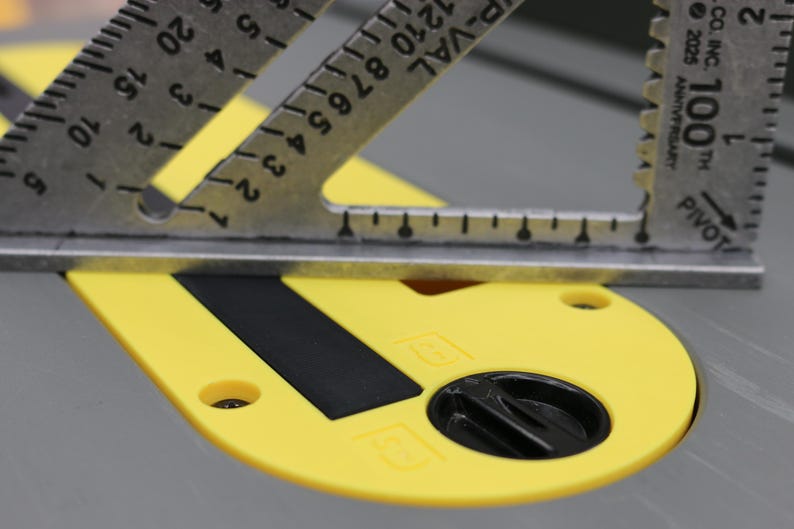 May include: A silver metal rafter square with black markings rests on a yellow circular table saw insert plate. The plate features a black rectangular slot and a black circular locking knob. The square displays the text 100th and PIVOT.