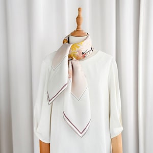 May include: A square silk scarf with a floral design in shades of yellow, pink, and orange. The scarf has a white background with a burgundy border. The scarf is draped over a white blouse on a wooden mannequin.