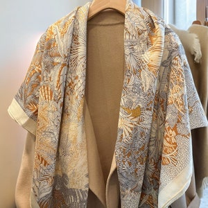 May include: A beige coat with a patterned silk scarf draped over it. The scarf features a botanical design in shades of gold, white, and gray. The coat is hanging on a wooden hanger, with another coat visible in the background.