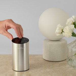 以下が含まれることがあります： 上部にコントロールパネルが付いたシルバーとブラックのエッセンシャルオイルディフューザー。ディフューザーには「OviPod」とラベルが貼られています。