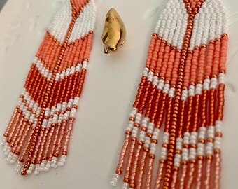 May include: A pair of long, beaded earrings with a geometric pattern in shades of coral, white, and copper. The earrings are made with small, round beads and have a fringe-like design at the bottom.