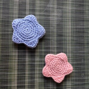 May include: Two crocheted star-shaped appliques, one in a light periwinkle blue and one in a soft pastel pink. Each piece features a dense, circular yarn stitch pattern radiating from the center to five rounded points, creating a textured finish.