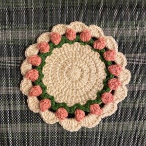 Könnte beinhalten: Ein gehäkelter Untersetzer mit einem beigefarbenen Zentrum und einem Rand aus rosa und grünem Garn. Der Rand besteht aus kleinen, gehäkelten Blüten.