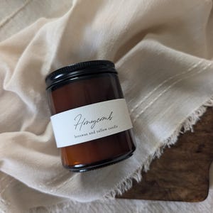 May include: A small amber glass jar candle with a black screw-top lid. A white paper label wraps around the centre, featuring the black printed text Honeycomb and beeswax and tallow candle in a simple, clean, and legible font style.