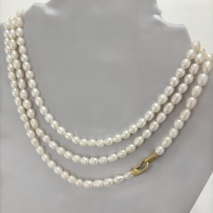 Peut inclure: Un collier de perles à trois rangs avec un fermoir doré. Les perles sont blanches et de forme ovale. Le collier est présenté sur une surface blanche.