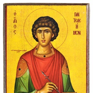May include: Icon depicting a saint with a halo, dressed in a red robe over a green garment. The saint holds a writing instrument and a small, gold-coloured box. The background is yellow with Greek text.