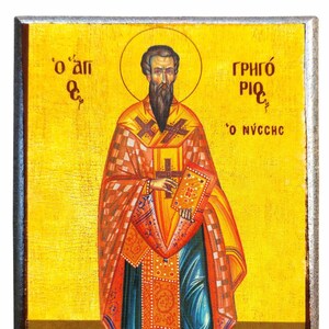 Op de afbeelding: Houten icoon met een religieuze figuur met een aureool, gekleed in sierlijke gewaden van rood, oranje en goud. De figuur houdt een boek vast en staat tegen een gouden achtergrond met Griekse tekst. Het icoon is omlijst in donker hout.