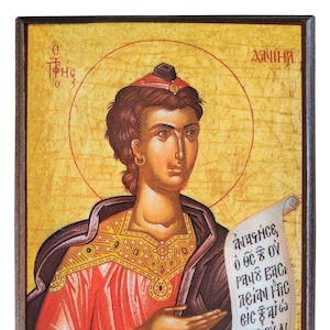 May include: A religious icon painting of a woman holding a scroll with Greek text. The woman is wearing a red and purple robe and a gold crown. The background is a yellow and gold color. The text on the scroll reads "Θαλασσε, ο ος ε ου ραυς βασι λεται ητις εις ξαιης μας ου αι αφοαρ η σεται."