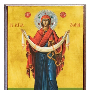 Puede incluir: Un icono religioso que representa a una mujer con una túnica roja y azul sosteniendo una banda blanca. El icono tiene un fondo dorado con texto griego que dice "Η ΑΓΙΑ ΖΩΝΗ".