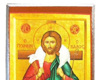 May include: Religious icon depicting a figure in red and green robes, holding a lamb. The background is gold with Greek text and a cross. The icon is framed in silver and brown.