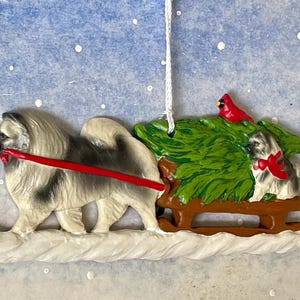 Puede incluir: Adorno navideño que representa un perro tirando de un trineo con otro perro y un cardenal. Los perros son grises y blancos, con detalles rojos. El trineo es marrón, sobre un fondo azul nevado.