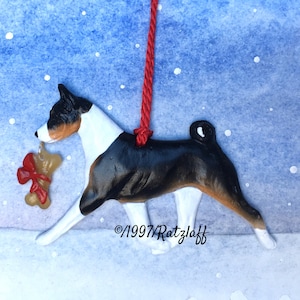 May include: A black, white, and brown Basenji dog ornament holding a red and brown bow-tied bone. The ornament is hanging from a red string against a blue and white snowy background. ©1997/Ratzlaff