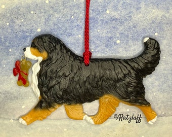 Può includere: Un ornamento a forma di bovaro del bernese con pelo nero, bianco e marrone, che tiene in bocca un ciondolo a forma di osso dorato con un fiocco rosso. L'ornamento è appeso a un cordino rosso e riporta la scritta Ratzlaff in nero.