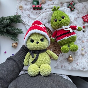 May include: Two crocheted Grinch dolls. One doll is wearing a red and white striped hat and a gray scarf. The other doll is wearing a red and black striped sweater and a white scarf. Both dolls are green with black eyes.