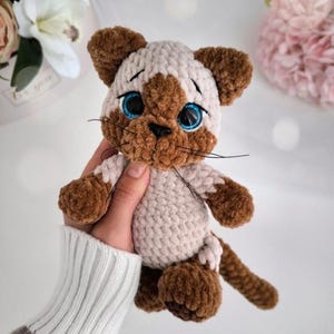 May include: A handmade crochet cat doll with brown and cream-colored yarn. The cat has blue eyes, a brown nose, and whiskers. The doll is being held in a person's hand. The cat doll is a cute and cuddly toy.