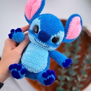 Puede incluir: Un peluche de ganchillo azul y rosa hecho a mano, con orejas grandes, interiores rosas y ojos negros grandes. El juguete tiene una barriga azul claro y patas azul oscuro, que se asemejan a un personaje de dibujos animados. El juguete est&aacute; hecho de hilo suave.