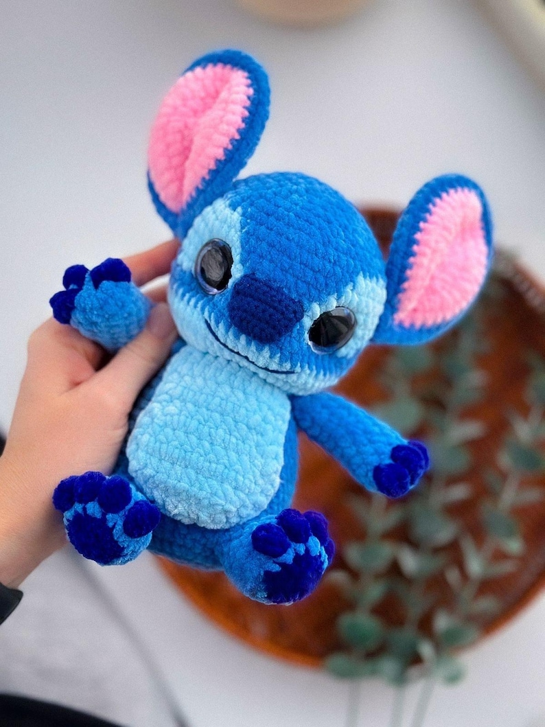Puede incluir: Un peluche de ganchillo azul y rosa hecho a mano, con orejas grandes, interiores rosas y ojos negros grandes. El juguete tiene una barriga azul claro y patas azul oscuro, que se asemejan a un personaje de dibujos animados. El juguete est&aacute; hecho de hilo suave.
