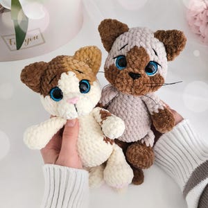 Peut inclure: Deux peluches chats crochetées à la main. L'un est marron et blanc, l'autre marron et beige. Les deux ont des yeux bleus et des nez roses. Les peluches sont tenues par une personne portant un pull rayé blanc et gris.