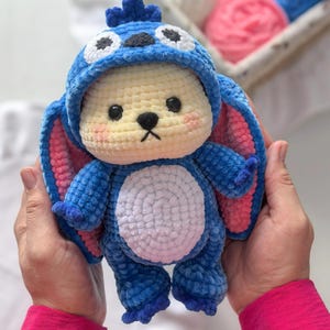 Peut inclure: Une peluche au crochet bleue et jaune faite &agrave; la main, ressemblant &agrave; un personnage de dessin anim&eacute;. La peluche a de grandes oreilles avec des d&eacute;tails roses &agrave; l'int&eacute;rieur, des yeux noirs et un ventre blanc. Le jouet est tenu dans deux mains.