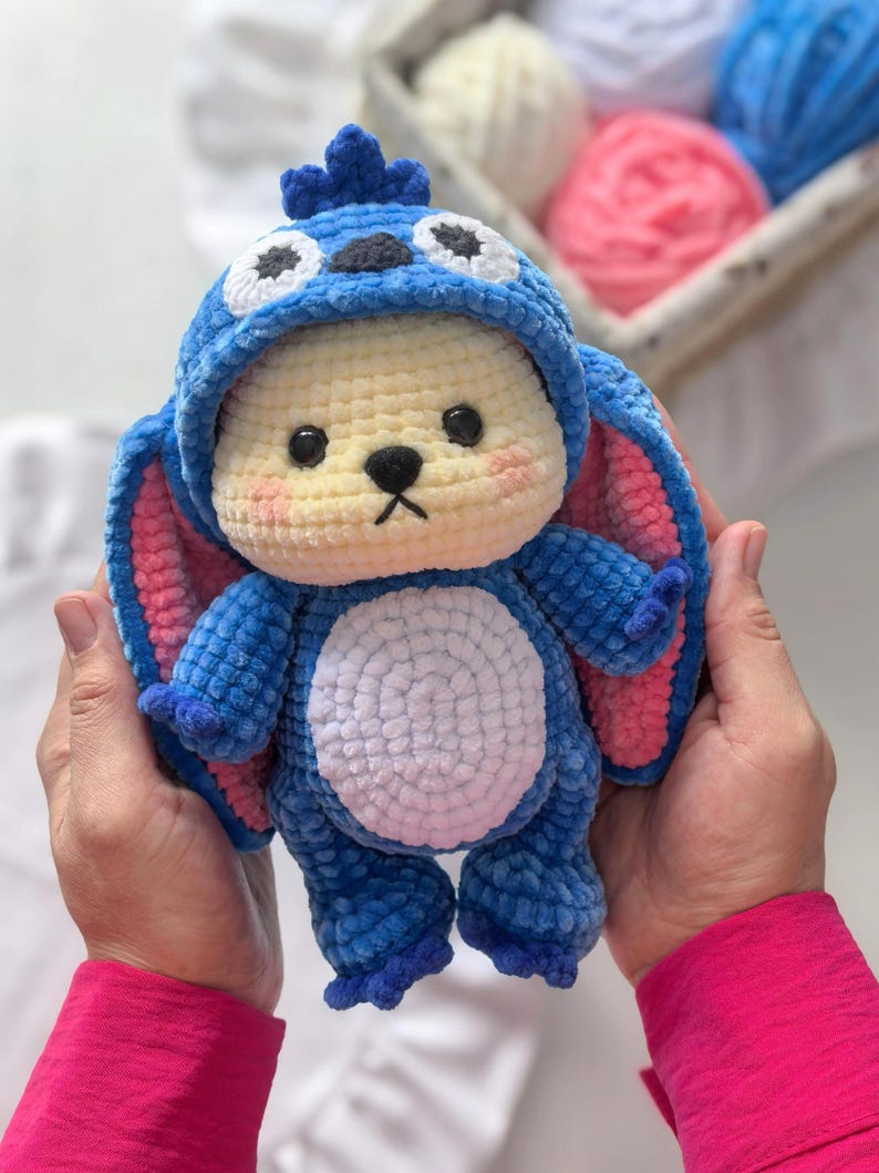 Peut inclure: Une peluche au crochet bleue et jaune faite &agrave; la main, ressemblant &agrave; un personnage de dessin anim&eacute;. La peluche a de grandes oreilles avec des d&eacute;tails roses &agrave; l'int&eacute;rieur, des yeux noirs et un ventre blanc. Le jouet est tenu dans deux mains.