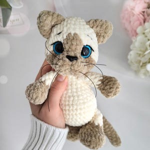 May include: A handmade Siamese cat plush toy. The cat is made of soft, cream-colored and brown yarn. It has blue eyes, black whiskers, and a small black nose. The plush cat is a cuddly, handcrafted item.
