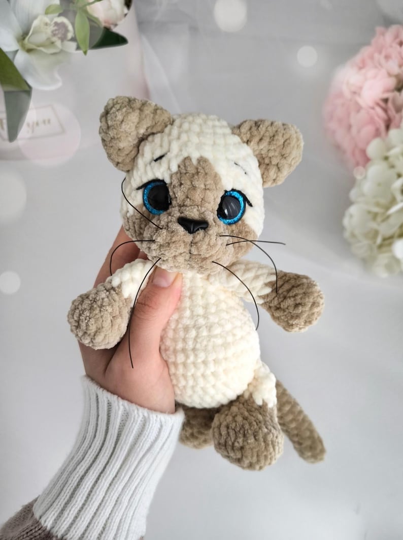May include: A handmade Siamese cat plush toy. The cat is made of soft, cream-colored and brown yarn. It has blue eyes, black whiskers, and a small black nose. The plush cat is a cuddly, handcrafted item.