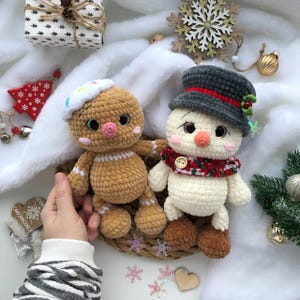 Può includere: Due peluche natalizi fatti a mano all'uncinetto: un omino di pan di zenzero con un cappello di glassa bianca e un pupazzo di neve con un cappello grigio e una sciarpa rossa. I giocattoli sono appoggiati su un cestino intrecciato con uno sfondo bianco soffice e decorazioni natalizie.