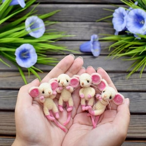 May include: Four handmade crochet mice with cream bodies, pink ears, feet, and tails are held in open hands. The mice are arranged in a row. The background includes blue flowers and green foliage.