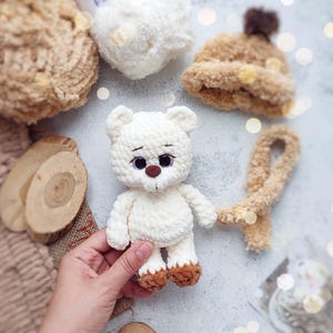 May include: A handmade white teddy bear with brown accents on the feet and nose. The bear is surrounded by other crocheted items, including a hat and scarf, all in neutral tones. The bear is approximately 6 inches tall.