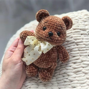 Peut inclure: Un ours en peluche marron crochet&eacute; avec des yeux en boutons noirs et un ruban cr&egrave;me nou&eacute; autour du cou. Le ruban est orn&eacute; de petits c&oelig;urs dor&eacute;s. L'ours est doux et moelleux.