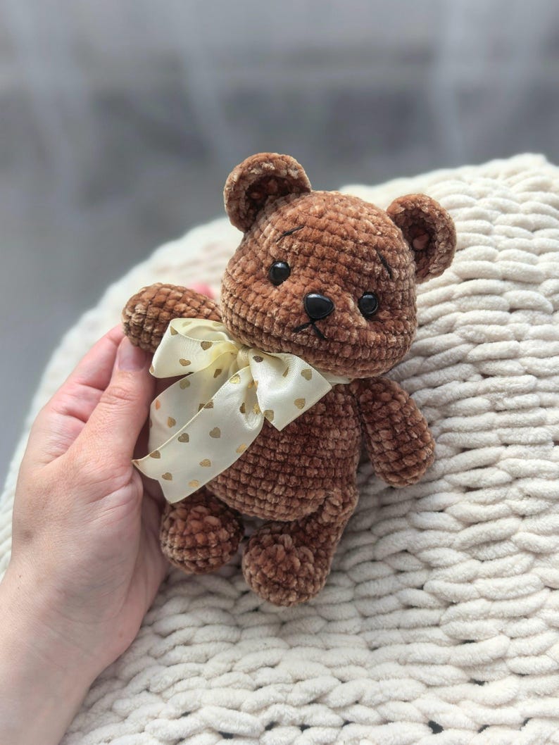 Peut inclure: Un ours en peluche marron crochet&eacute; avec des yeux en boutons noirs et un ruban cr&egrave;me nou&eacute; autour du cou. Le ruban est orn&eacute; de petits c&oelig;urs dor&eacute;s. L'ours est doux et moelleux.