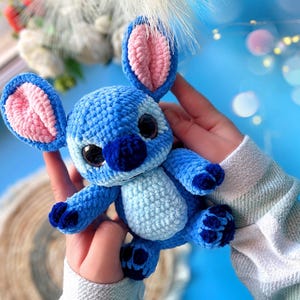 Puede incluir: Un peluche de ganchillo azul hecho a mano con orejas grandes, una nariz azul oscuro y ojos negros grandes. El interior de las orejas es rosa. El juguete se sostiene en las manos de una persona sobre un fondo azul claro.
