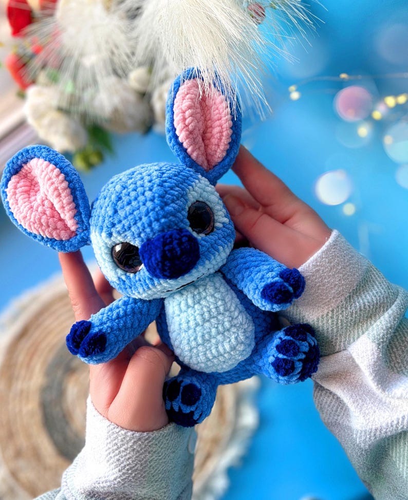 Puede incluir: Un peluche de ganchillo azul hecho a mano con orejas grandes, una nariz azul oscuro y ojos negros grandes. El interior de las orejas es rosa. El juguete se sostiene en las manos de una persona sobre un fondo azul claro.