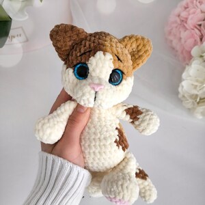 May include: A handmade crochet cat plushie in white, brown, and tan. The cat has blue eyes, a pink nose, and brown spots. The plushie is held in a hand, showcasing its soft texture and detailed craftsmanship.