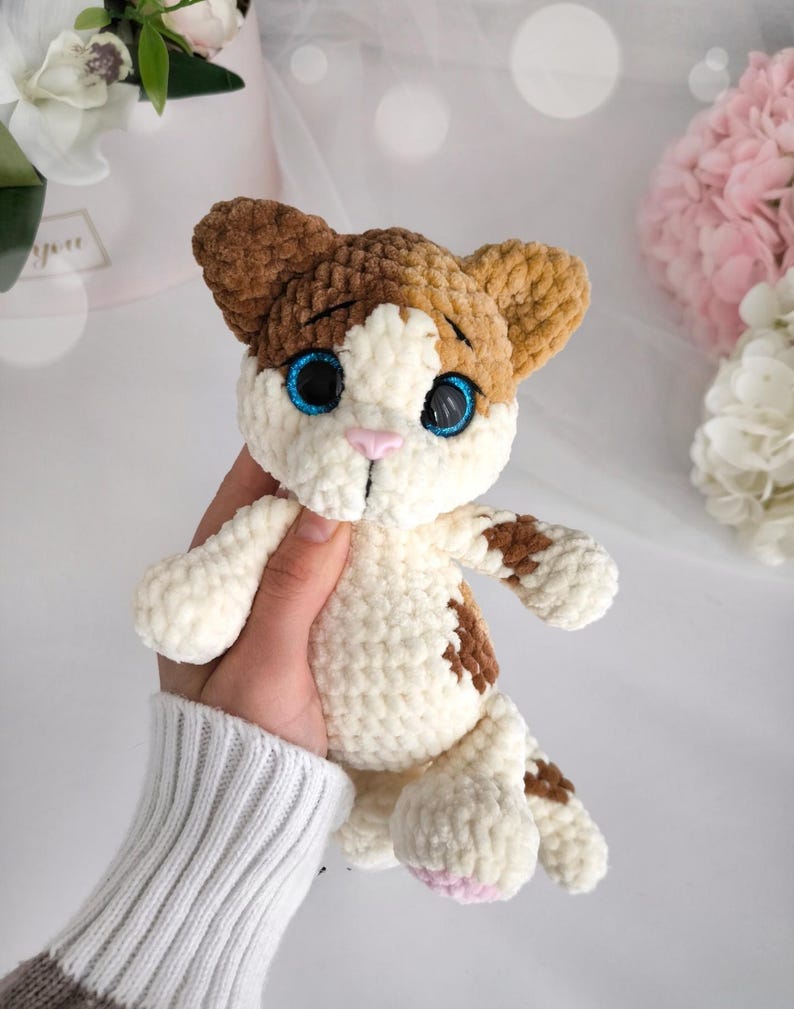 May include: A handmade crochet cat plushie in white, brown, and tan. The cat has blue eyes, a pink nose, and brown spots. The plushie is held in a hand, showcasing its soft texture and detailed craftsmanship.