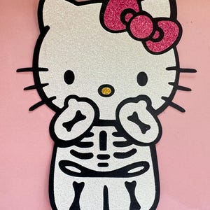 May include: A glittery white Hello Kitty-shaped decoration with a pink glitter bow and black outline. The character is dressed as a skeleton, with a black and white skeleton design on the body and arms. The background is pink.