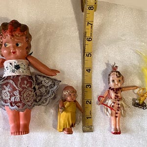 May include: Three vintage dolls are displayed, each with unique outfits and features. The largest doll wears a lace skirt and a top with a floral design. A measuring tape is next to the dolls. The smallest doll is wearing a yellow dress.