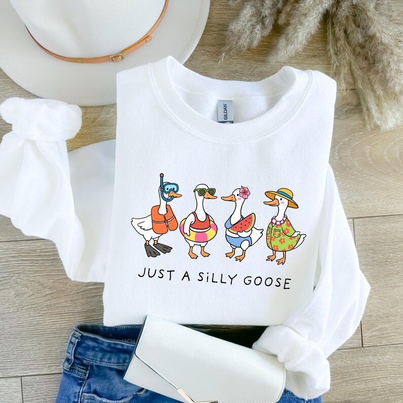 May include: White sweatshirt with a graphic of four cartoon geese in summer attire, including a life vest, sunglasses, a floral swim ring, and a hat. The text "JUST A SILLY GOOSE" is printed below the geese.