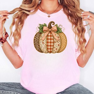 May include: A light pink t-shirt featuring a central graphic of a pumpkin. The pumpkin is divided into vertical sections with leopard print, floral patterns, brown and white gingham, and polka dots, topped with a tan bow and green leaves.